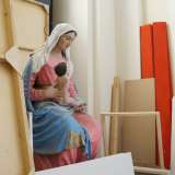 Una statua della Vergine col bambino in abiti rosa e azzurro, giace invece silenziosa dietro tele e cornici di legno 